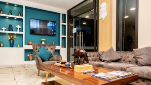 A modern living room with a large flat-screen TV, a brown leather sofa, a wooden coffee table, and teal shelving with decorative items. - HOTEL GOLDEN PLAZZA CONAKRY (Conakry)