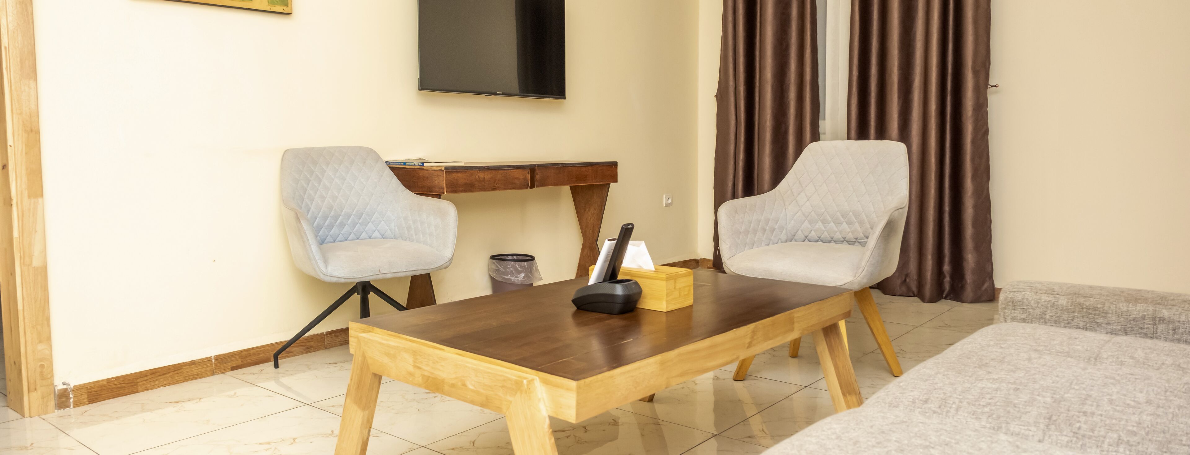 Habitación de hotel con una mesita de café de madera, dos sillones tapizados de gris, un pequeño escritorio de madera, un televisor montado en la pared y una ventana con cortinas marrones.