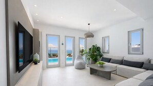 A modern living room with a large TV, a sofa, a coffee table, and a view of the pool and ocean. - Black Pearl Villa - Sea Front (Ampelas)