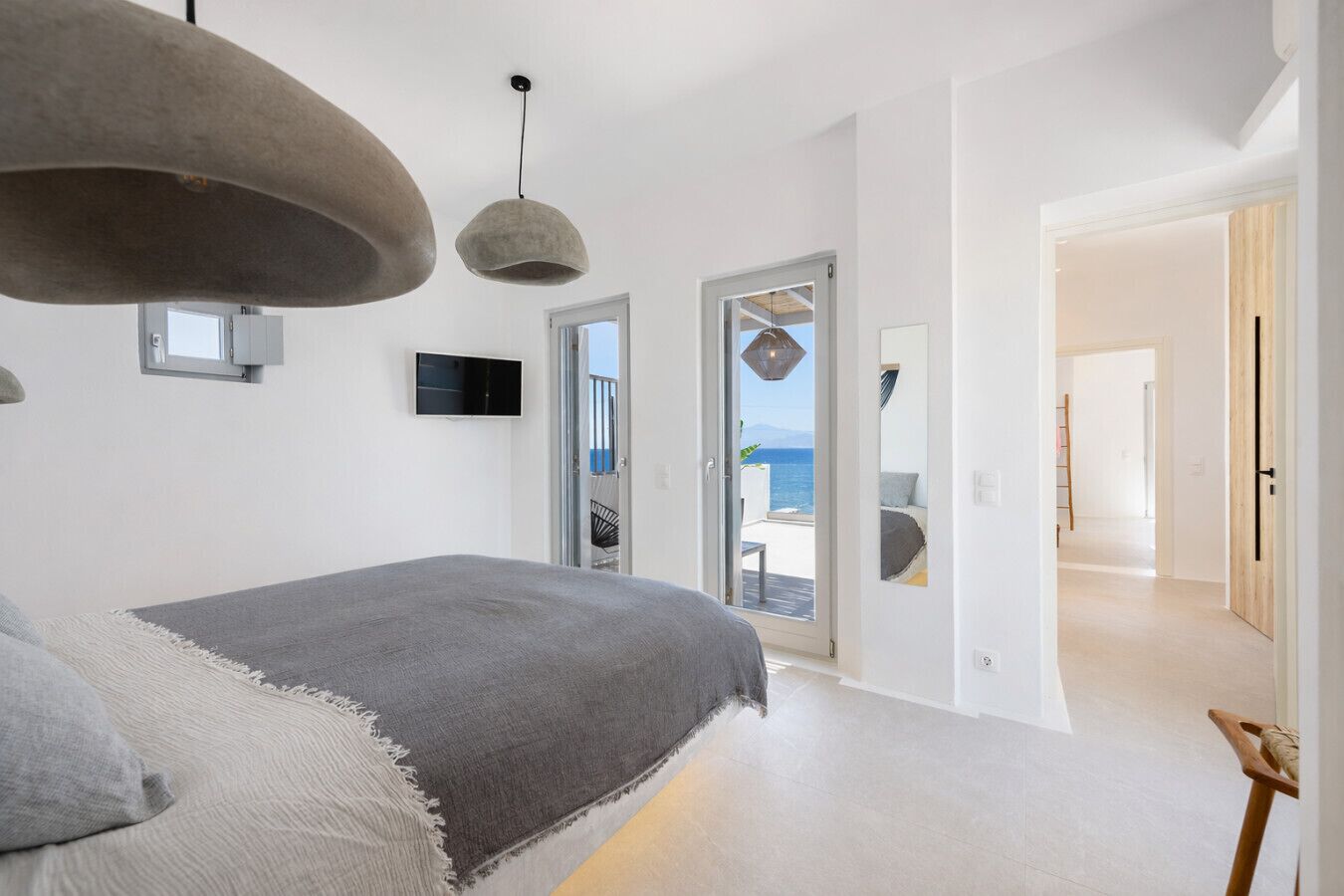 A modern bedroom with a large bed, a TV, and a balcony with a view of the sea.