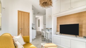 A modern living room with a sofa, a TV mounted on the wall, and a kitchen area in the background. - Folia House - Overlooking (Aspro Chorio)