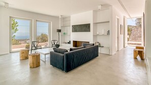 A spacious living area with a large sofa, a coffee table, and a fireplace. There are large windows offering a view of the outdoors. - Luxury standards in great location (Kostos)