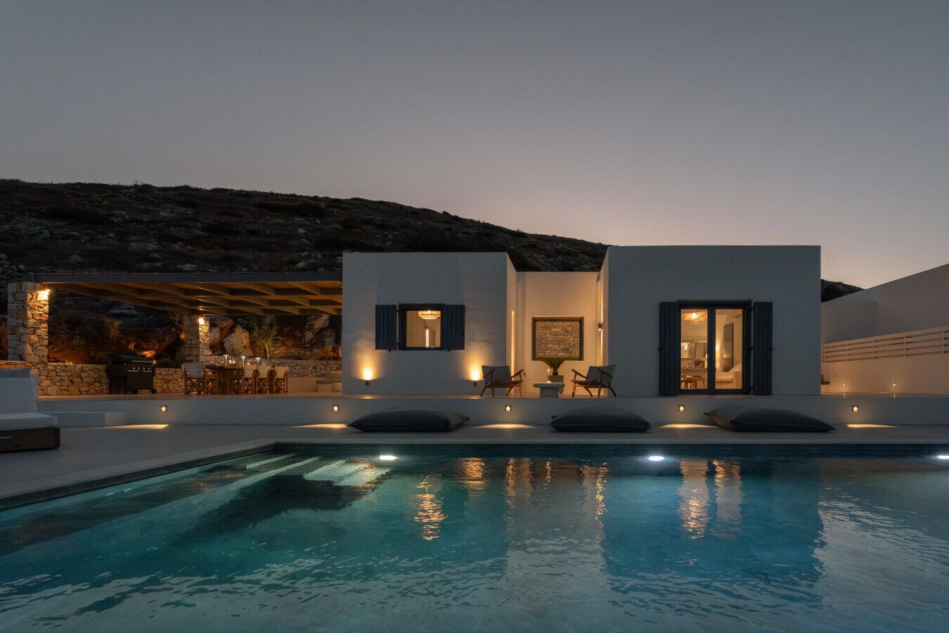 A modern outdoor pool area with a stone wall, two white buildings with windows, and a dining area with outdoor furniture.