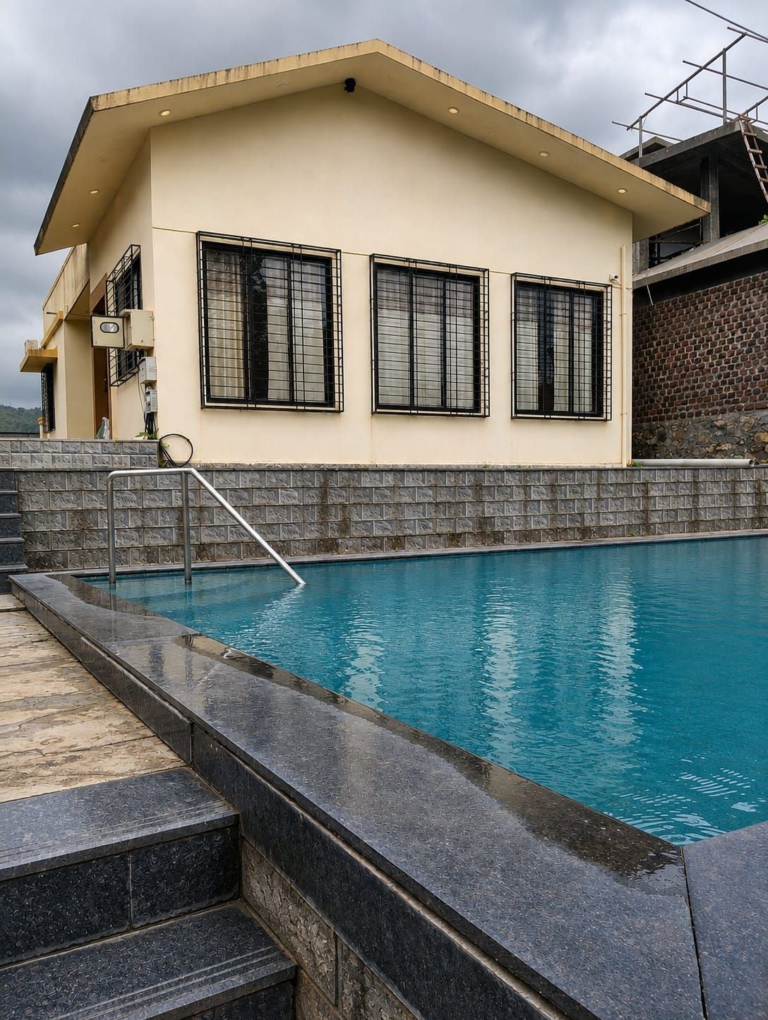Una piscina con borde de piedra, un edificio con fachada color crema y un muro de ladrillo.