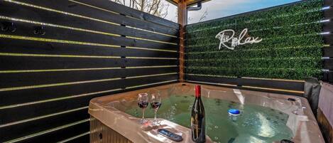 A hot tub with two glasses of red wine and a bottle, surrounded by wooden slats and a green hedge wall.