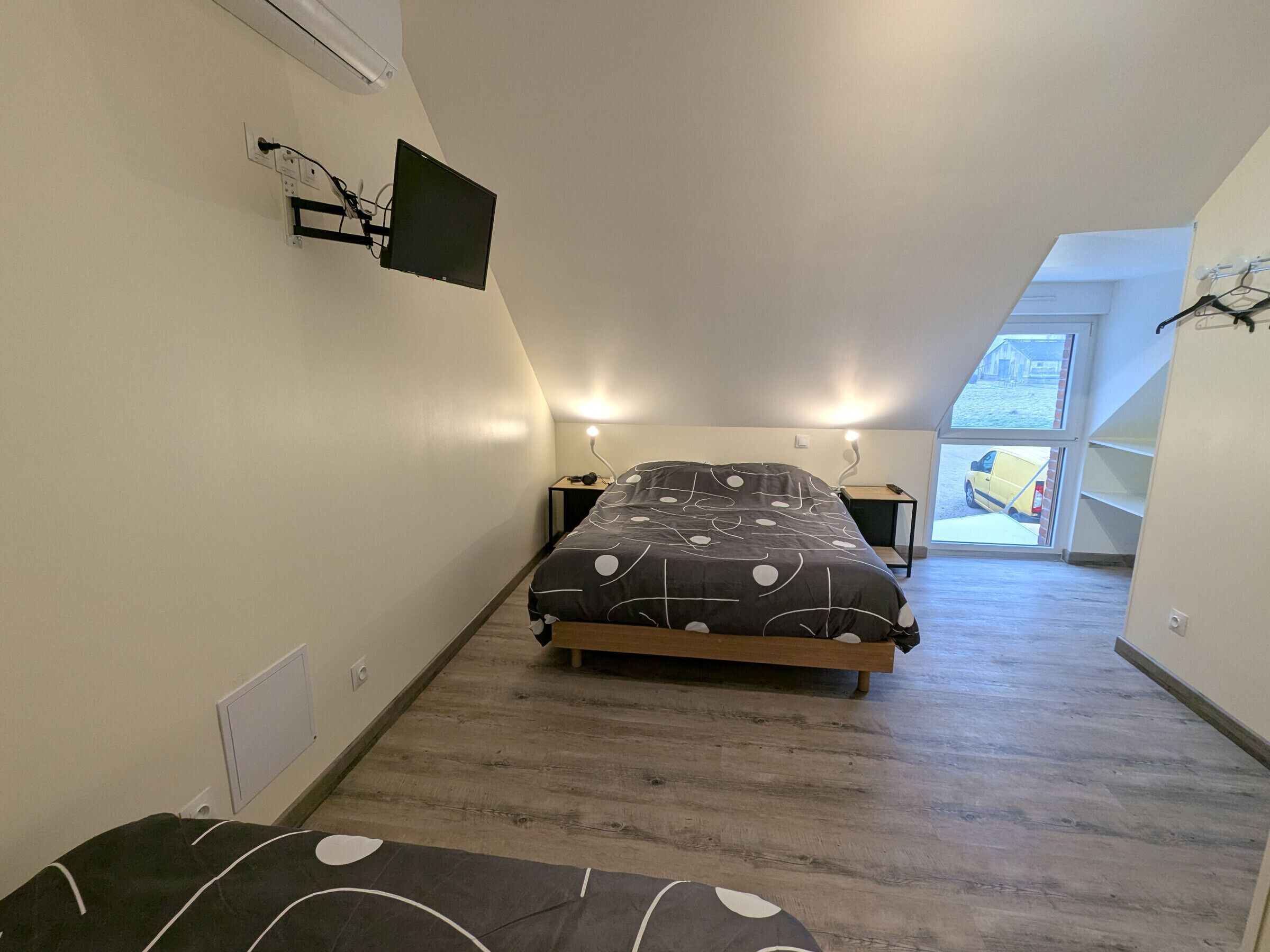 Un dormitorio con cama, mesitas de noche, un televisor de pantalla plana montado en la pared y una ventana que da a la calle, donde se ve una furgoneta amarilla.