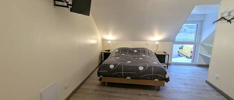 Un dormitorio con cama, mesitas de noche, un televisor de pantalla plana montado en la pared y una ventana que da a la calle, donde se ve una furgoneta amarilla.