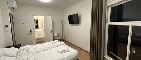 A hotel room with a bed, a TV mounted on the wall, a window with curtains, a coat rack, and a bathroom with a mirror and sink.