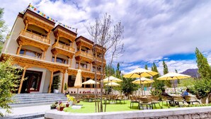 A multi-story building with balconies, outdoor seating with umbrellas, and a stone wall in the foreground. - Legacy Hotel Alchi (Alchi)