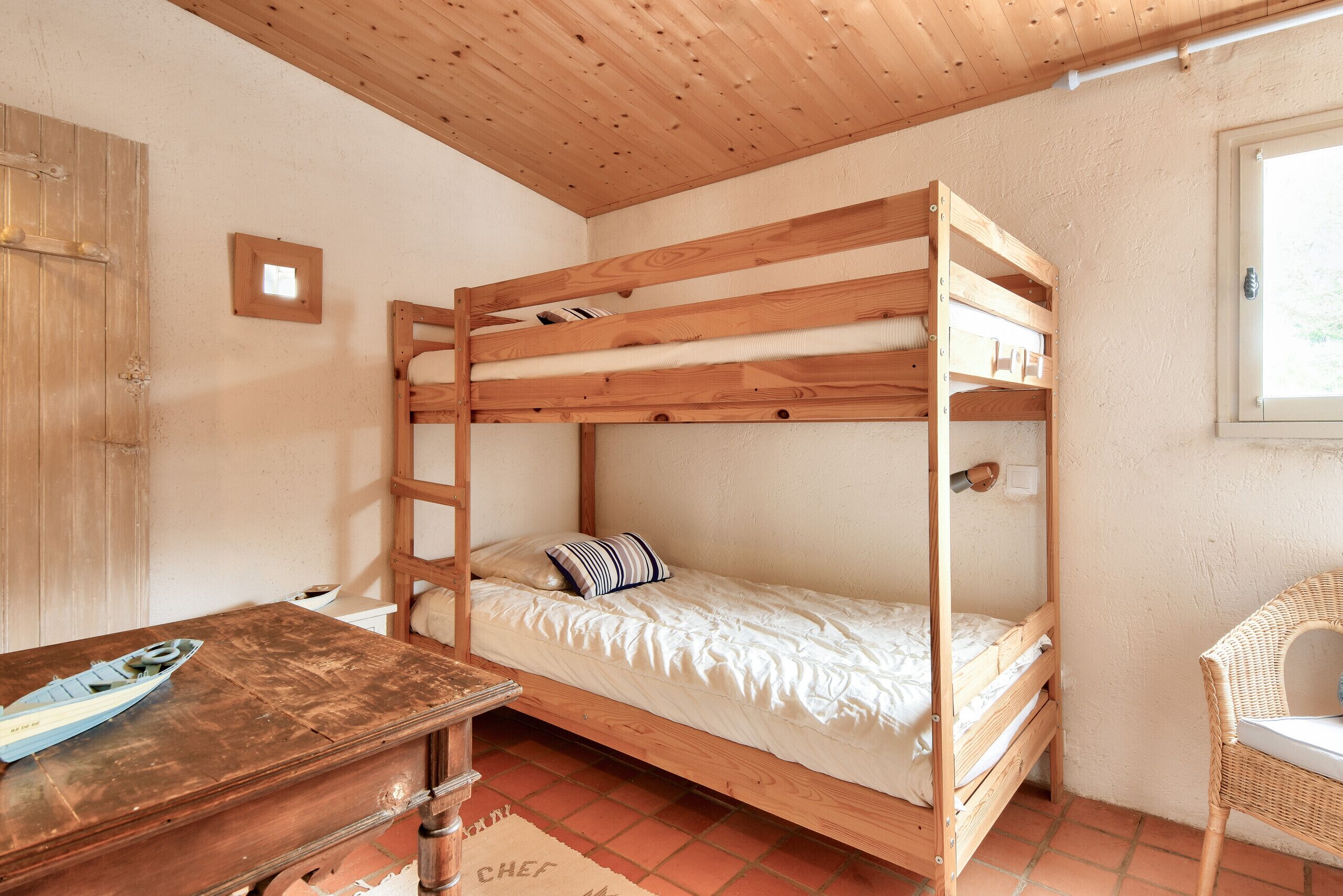 Una cama individual con estructura de madera y ropa de cama blanca, un escritorio de madera con un modelo de barco, una silla de mimbre y un pequeño cuadro en la pared.