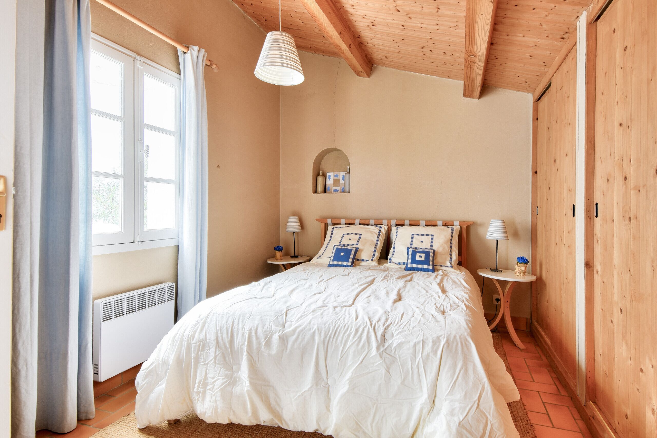 Een slaapkamer met een groot bed, twee nachtkastjes, een raam met gordijnen, een houten plafond en een kleine spiegel aan de muur.