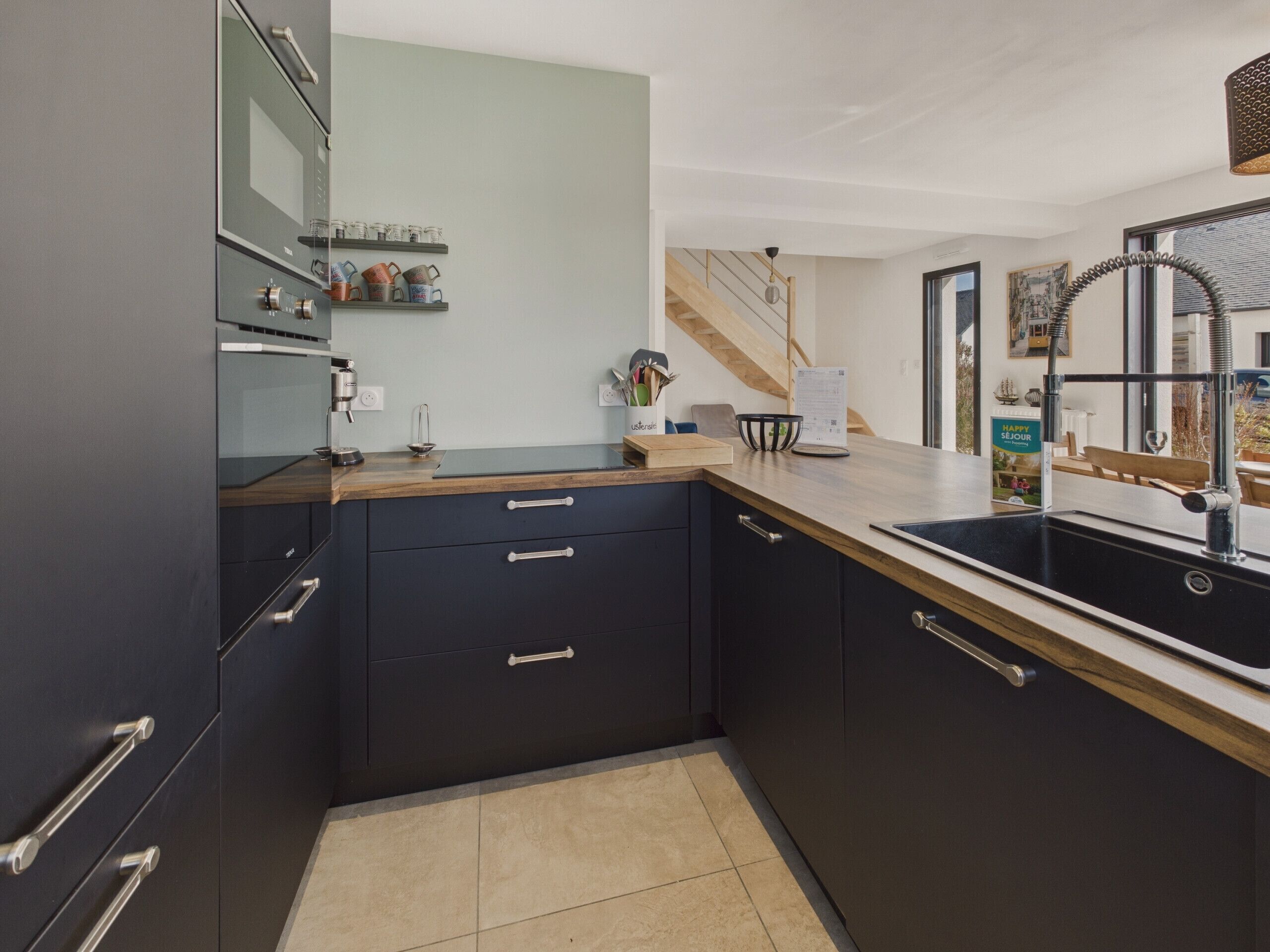 A modern kitchen with dark cabinetry, a wooden countertop, and a black sink. There is a small shelf with kitchen utensils and a bowl.