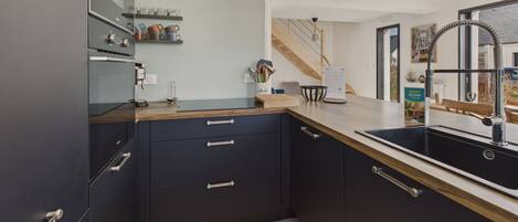A modern kitchen with dark cabinetry, a wooden countertop, and a black sink. There is a small shelf with kitchen utensils and a bowl.