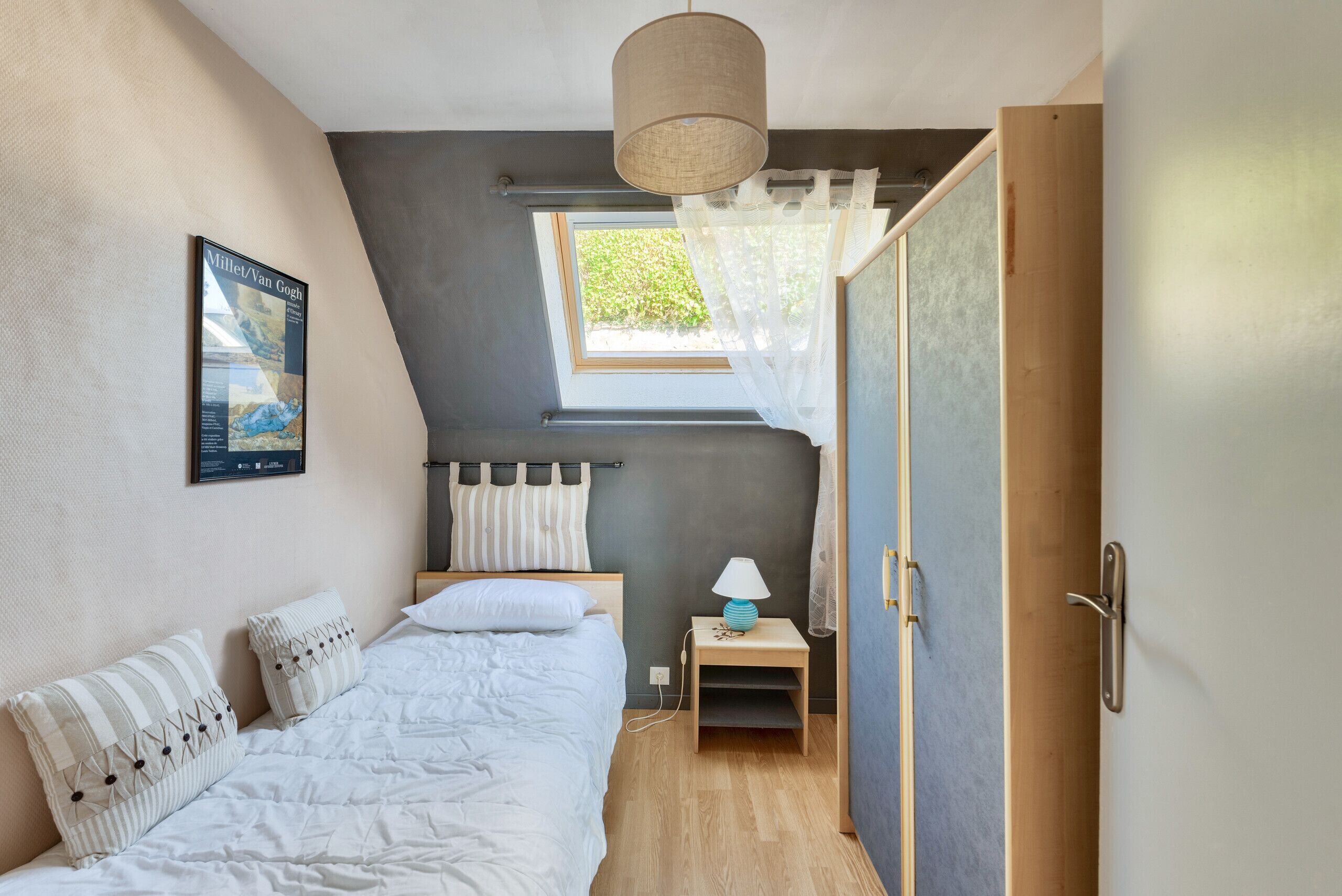 A bedroom with a bed, a nightstand with a lamp, a window with curtains, a door, and a wall-mounted picture.
