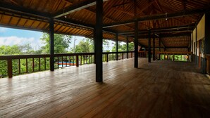 A spacious covered area with wooden flooring and railings, featuring a view of greenery and a swimming pool in the background. - Plengkung Lodge G-land (Kalipait)