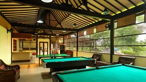 A pool hall with multiple pool tables, wooden benches, and large windows offering views of greenery. - Plengkung Lodge G-land (Kalipait)