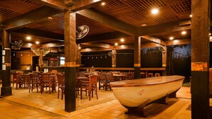 A restaurant with wooden tables and chairs, a large boat in the center, and a tiled floor. - Plengkung Lodge G-land (Kalipait)