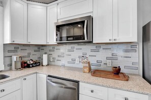 A kitchen with white cabinets, a stainless steel microwave, and a tiled backsplash. - Coastal Cottage - Upscale 2 BR Condo at Mini Golf (Topsail Beach)
