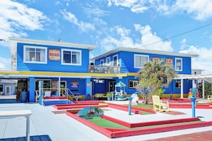 A two-story building with a blue and yellow facade, a small outdoor seating area, and a palm tree. - Coastal Cottage - Upscale 2 BR Condo at Mini Golf (Topsail Beach)