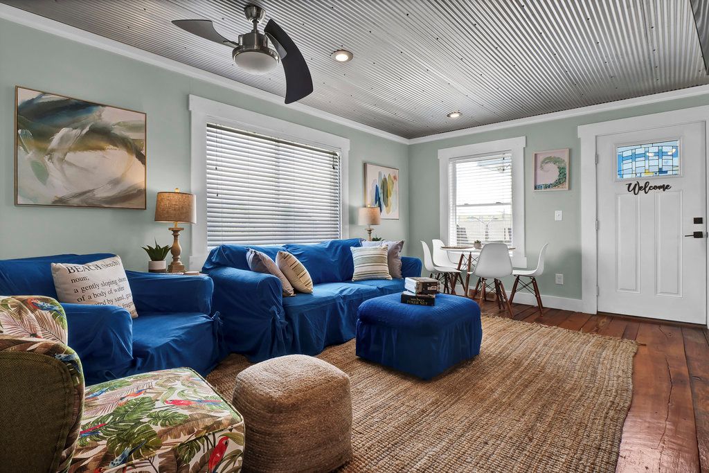 A living room with a blue sofa set, a coffee table, and a patterned armchair. There is a ceiling fan, a rug, and a door with a 'Welcome' sign.