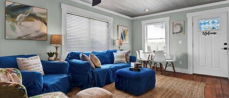 A living room with a blue sofa set, a coffee table, and a patterned armchair. There is a ceiling fan, a rug, and a door with a 'Welcome' sign.