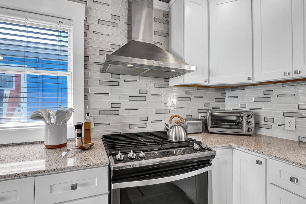 A modern kitchen with a gas stove, a stainless steel range hood, white cabinets, a toaster, and a window with blinds.