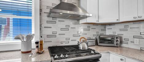 Una cocina moderna con estufa a gas, campana extractora de acero inoxidable, armarios blancos, un tostador y una ventana con persianas.