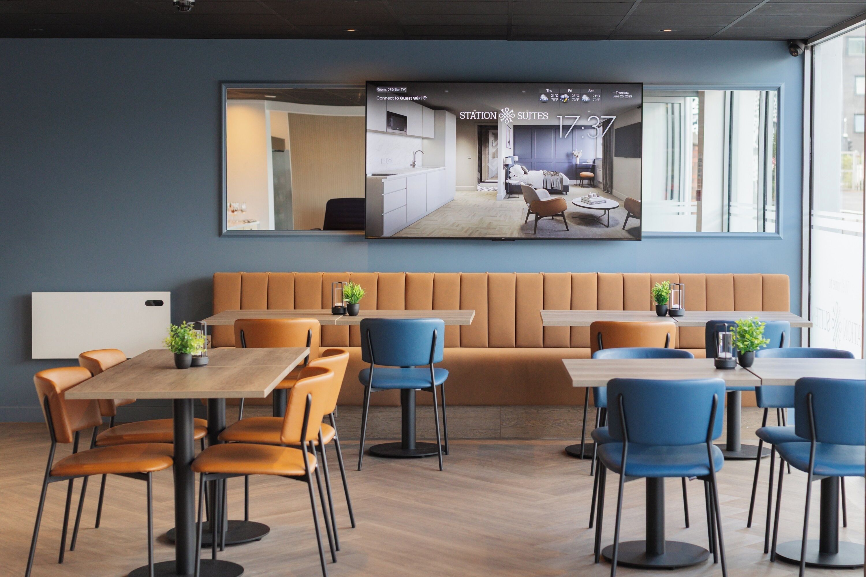 A modern hotel lobby with a large TV displaying a room layout, a seating area with orange and blue chairs, and a wooden floor.