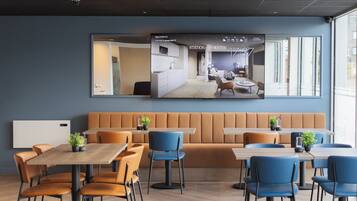 A modern hotel lobby with a large TV displaying a room layout, a seating area with orange and blue chairs, and a wooden floor.