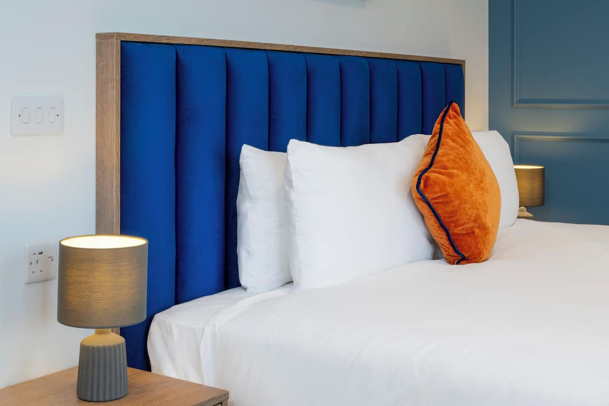A neatly made bed with white bedding and an orange pillow, a blue upholstered headboard, and a bedside lamp on a wooden nightstand.
