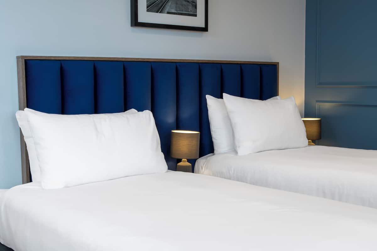 A hotel room with two beds, each with white bedding and a blue headboard. There are two bedside tables with lamps.