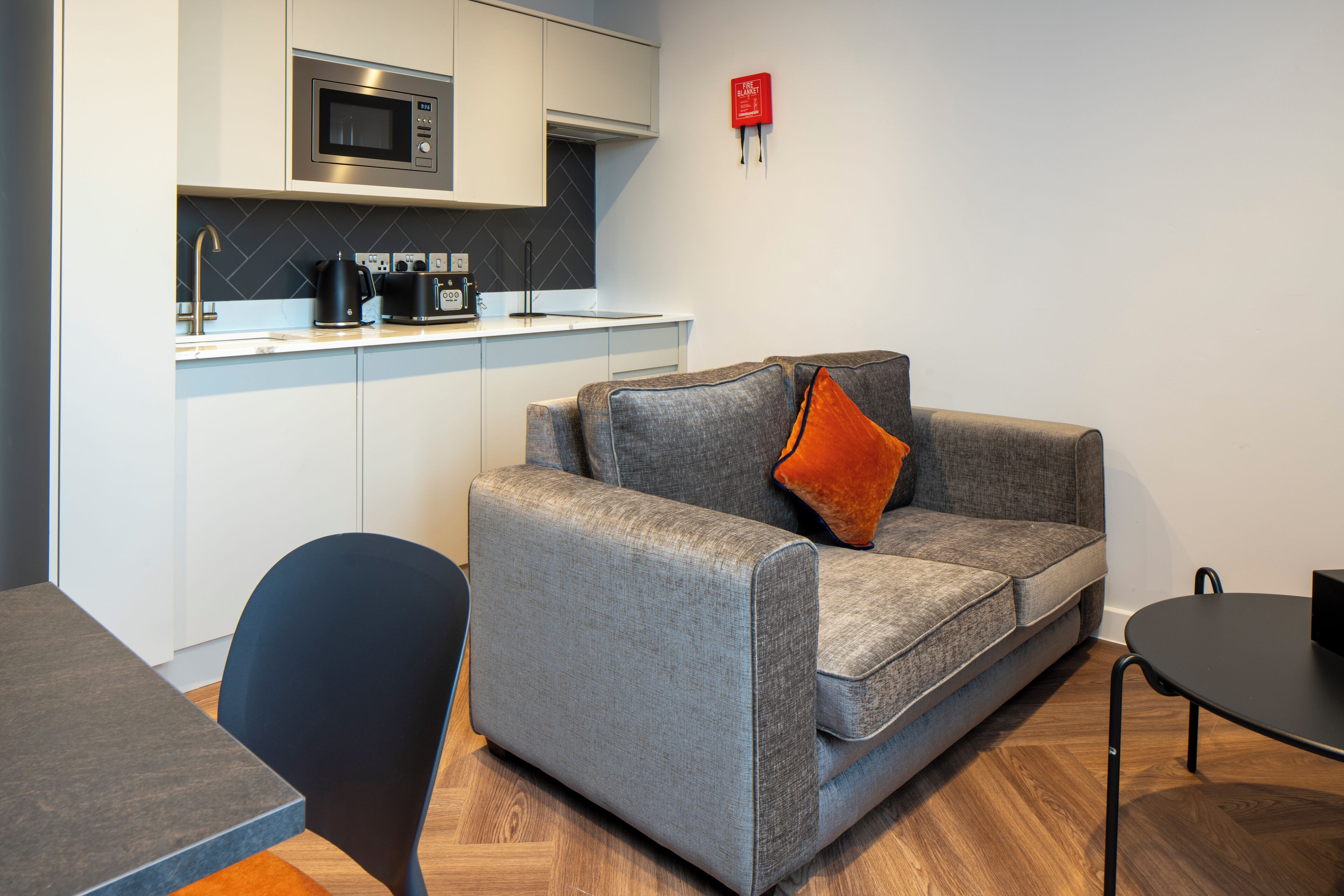 Een moderne kitchenette met een magnetron, koffiezetapparaat en een klein aanrecht. Er is een grijze bank met een oranje kussen en een zwarte stoel. Een klein tafeltje is rechts zichtbaar.