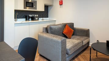 Een moderne kitchenette met een magnetron, koffiezetapparaat en een klein aanrecht. Er is een grijze bank met een oranje kussen en een zwarte stoel. Een klein tafeltje is rechts zichtbaar.
