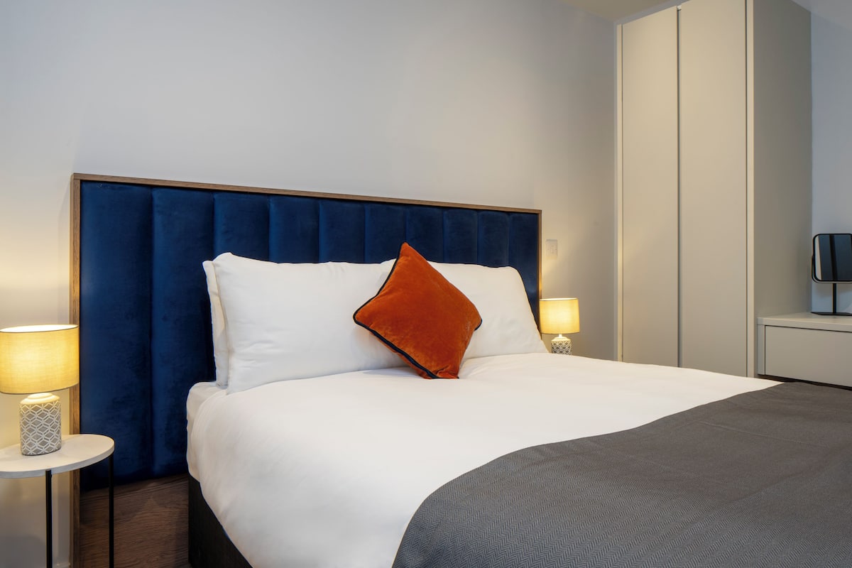 A neatly made bed with a blue headboard, white and orange pillows, and a grey throw blanket. There are two bedside tables with lamps.