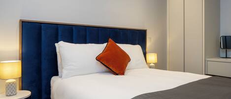 Una cama bien hecha con un cabecero azul, almohadas blancas y anaranjadas, y una manta gris. Hay dos mesitas de noche con lámparas.