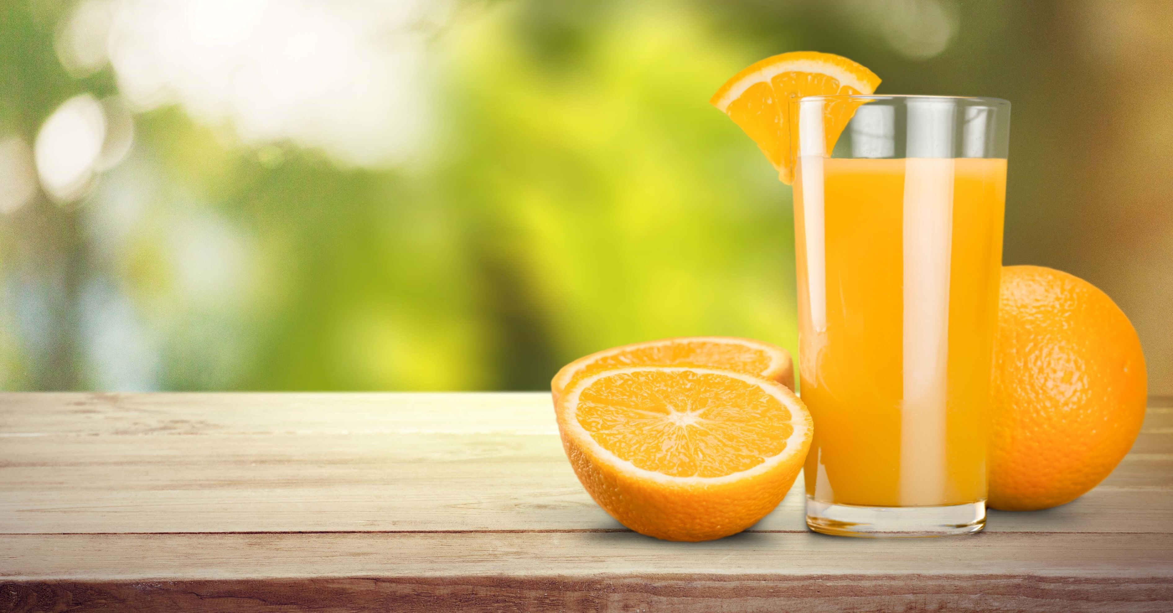 Een glas sinaasappelsap met een schijf sinaasappel op de rand, een gehalveerde sinaasappel en een hele sinaasappel op een houten ondergrond.