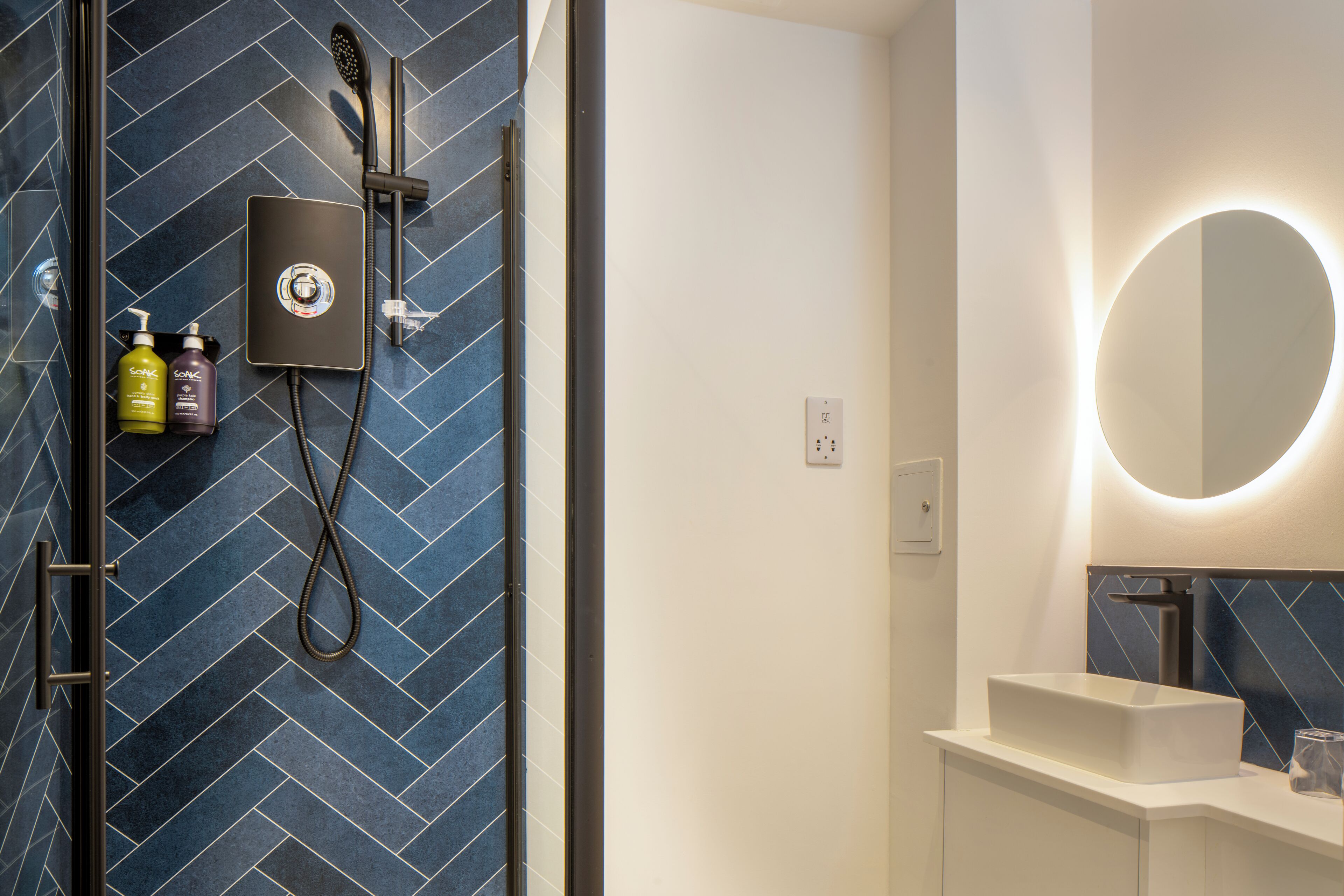 A modern bathroom with a shower, a handheld showerhead, a handheld dryer, and a large mirror with a circular light.