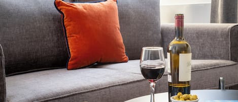 A grey sofa with a red pillow, a wine bottle, a glass of red wine, and a bowl of olives on a round table.