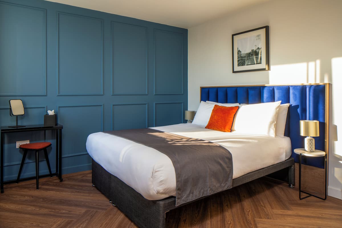 A neatly arranged hotel room with a large bed, a desk with a chair, and a small table with a lamp.