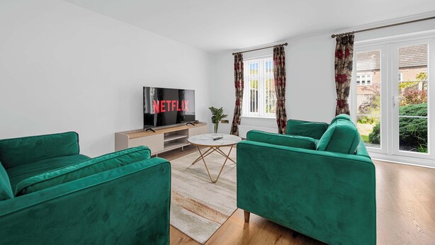 A modern living room with green sofas, a TV, and a view of a garden through large windows. - 3 Bed Pendlebury Home | Free Parking | Pets (Pendlebury)