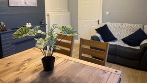 A dining area with a wooden table and chairs, a potted plant, a grey couch with patterned cushions, and a door in the background. - Welcoming 3-bedroom cottage with WiFi in Cork (Cork)