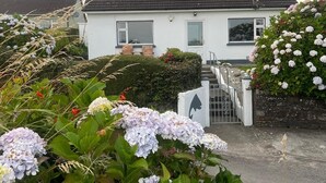 A white house with a dark roof, surrounded by lush greenery and hydrangea flowers. - Welcoming 3-bedroom cottage with WiFi in Cork (Cork)