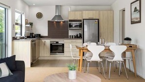 Una cocina moderna con horno empotrado, microondas y refrigerador de acero inoxidable. Hay un comedor con sillas blancas y una mesa de madera.