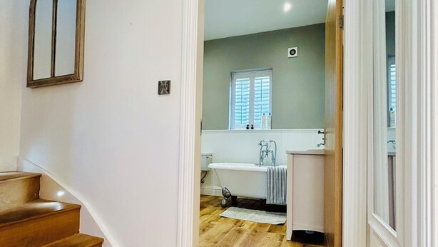 A hallway with wooden flooring leading to a bathroom with a bathtub and a window.