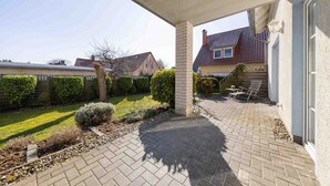 En overdækket terrasse med røde mursten, et træhegn og udsigt til et hus med rødt tag.
