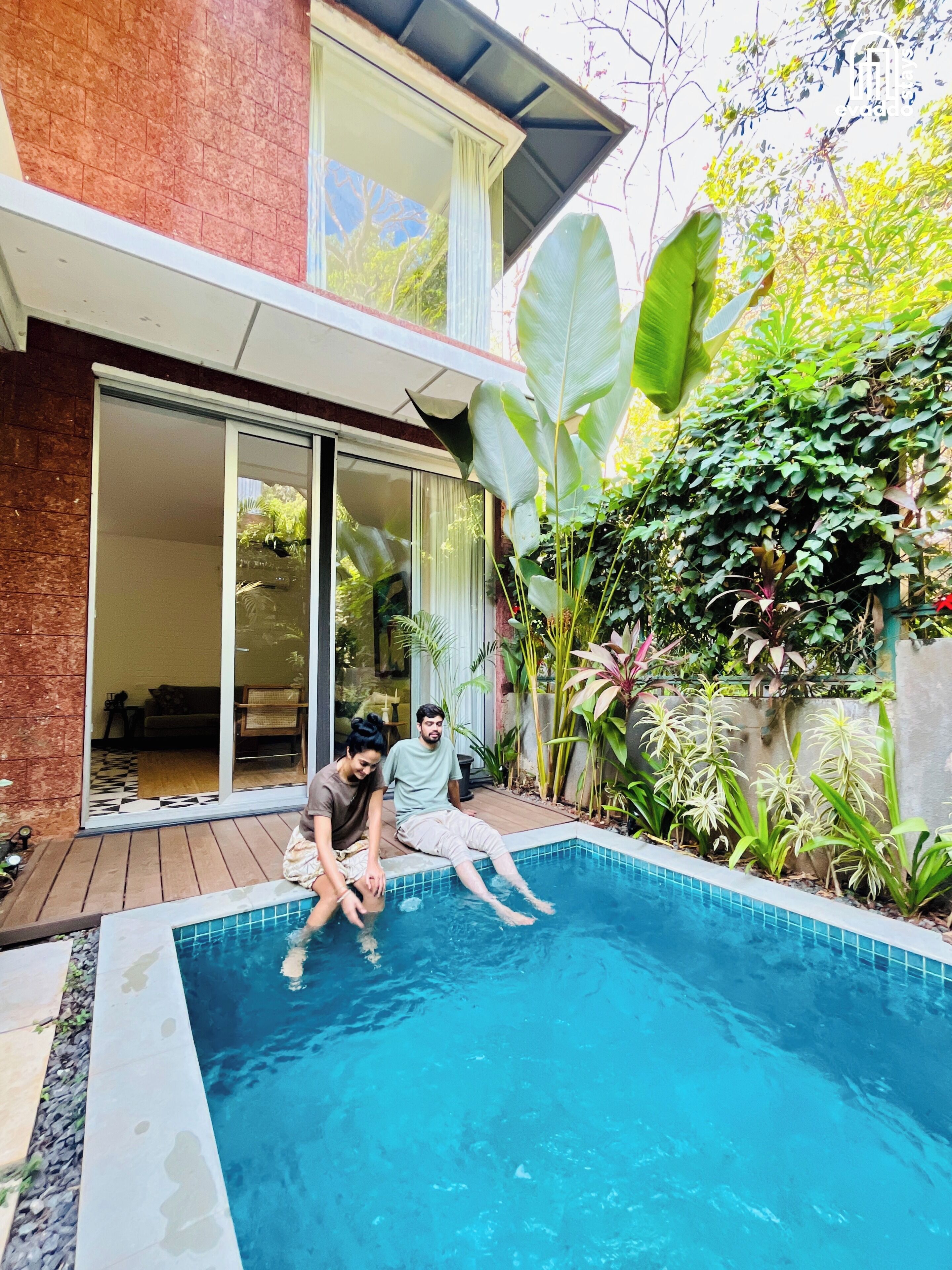 A swimming pool with two people sitting on the edge, surrounded by lush greenery and a modern house with large windows and a brick exterior.