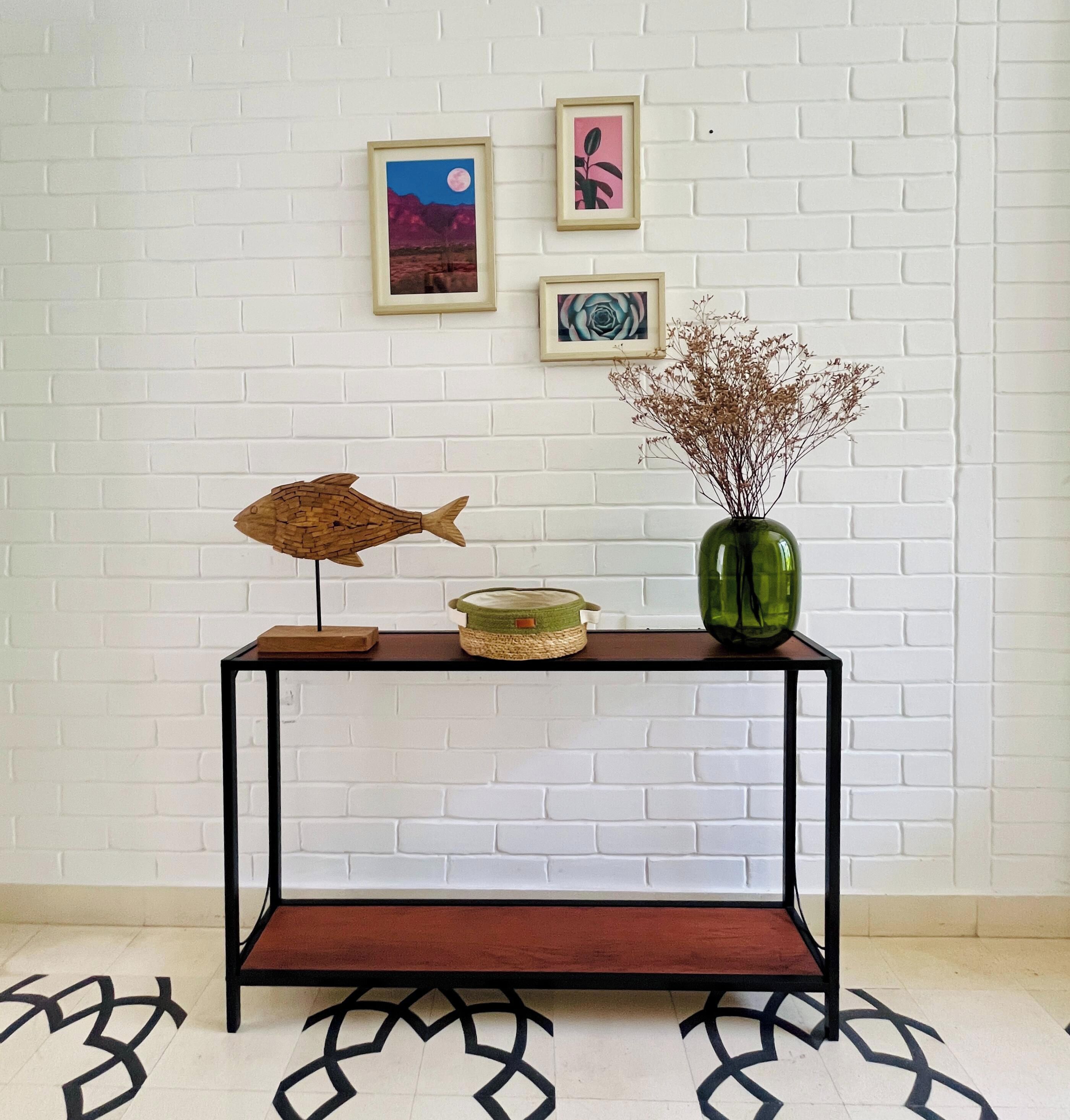 A console table with a fish sculpture, a vase with dried flowers, and framed artwork on a white brick wall.