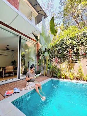 Una persona sentada junto a una piscina leyendo un libro, rodeada de vegetación y una casa moderna.