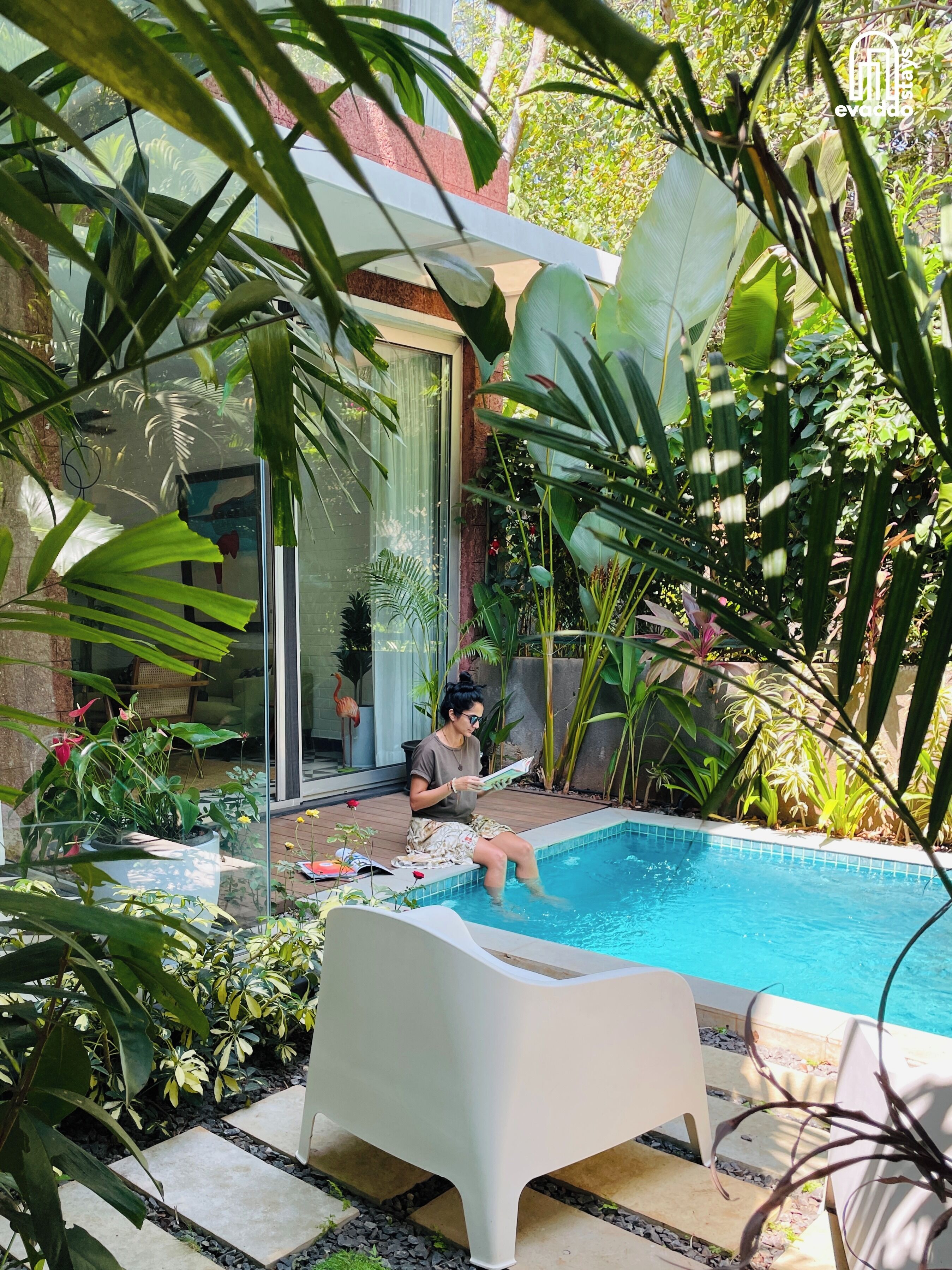 A person sitting by a pool with a book, surrounded by lush greenery and a modern house.