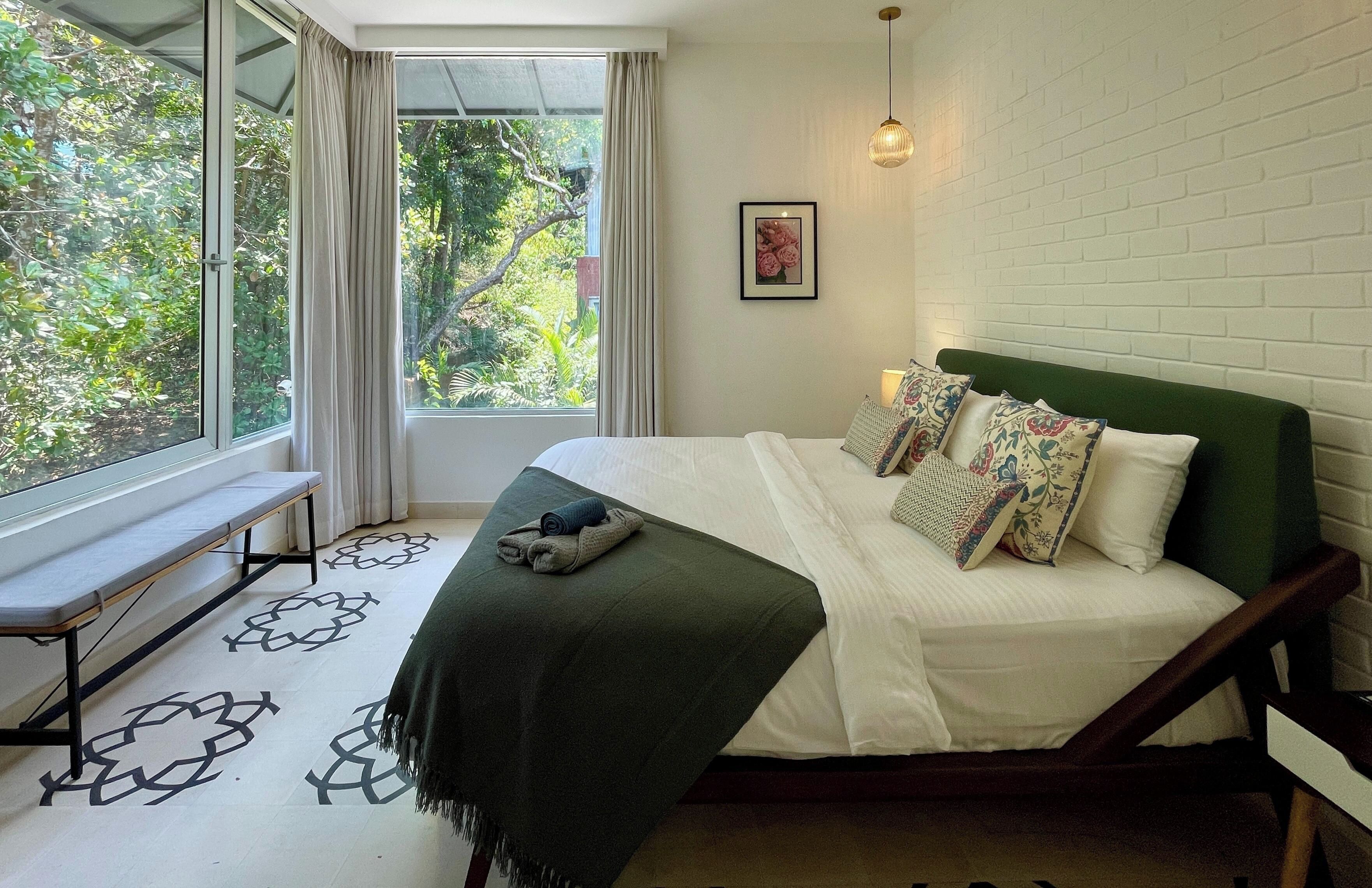 A bedroom with a bed, a bench, a window with curtains, and a view of greenery.